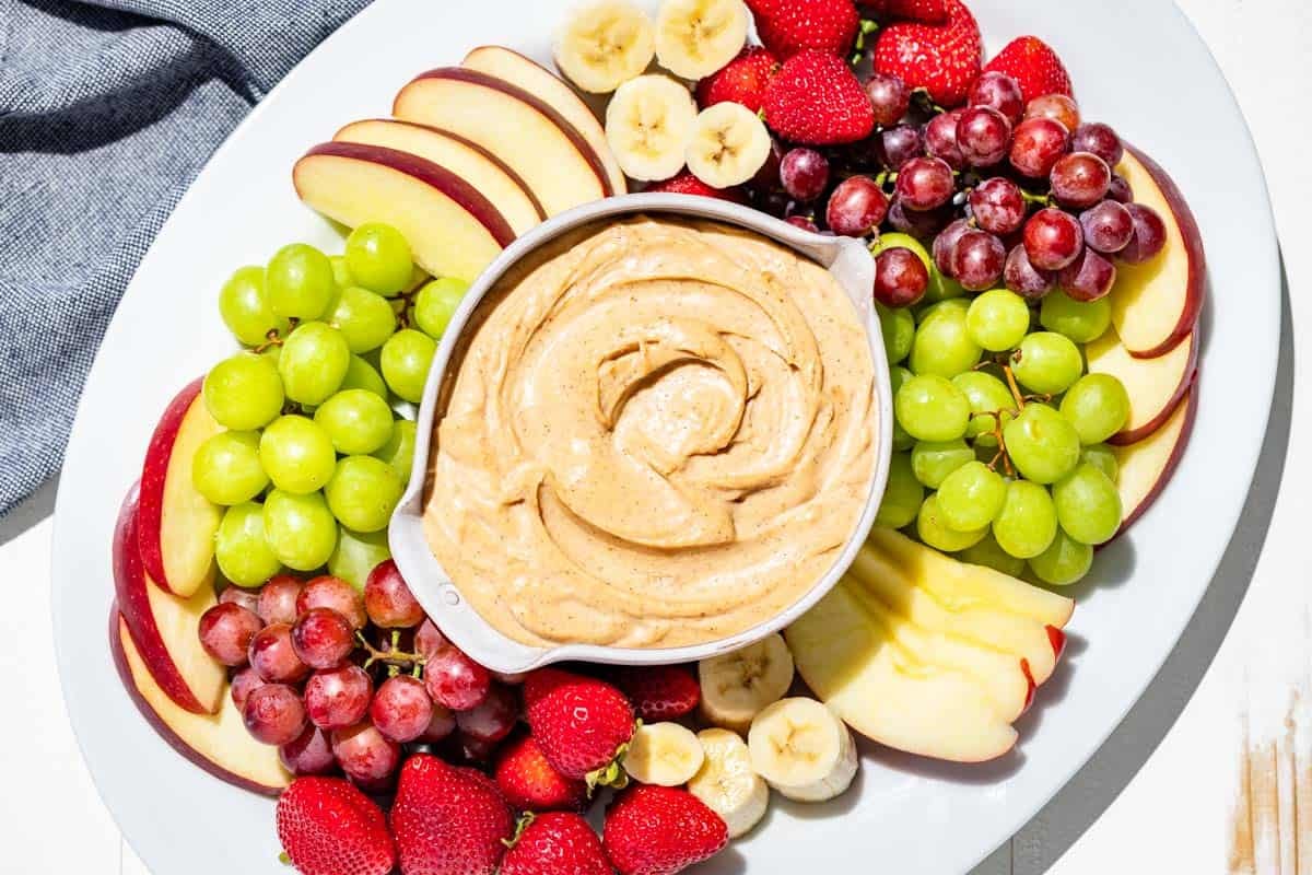 Finished peanut butter dip in a pottery bowl in the center of a large white platter surrounded by apple slices, grapes, strawberries, and banana slices.