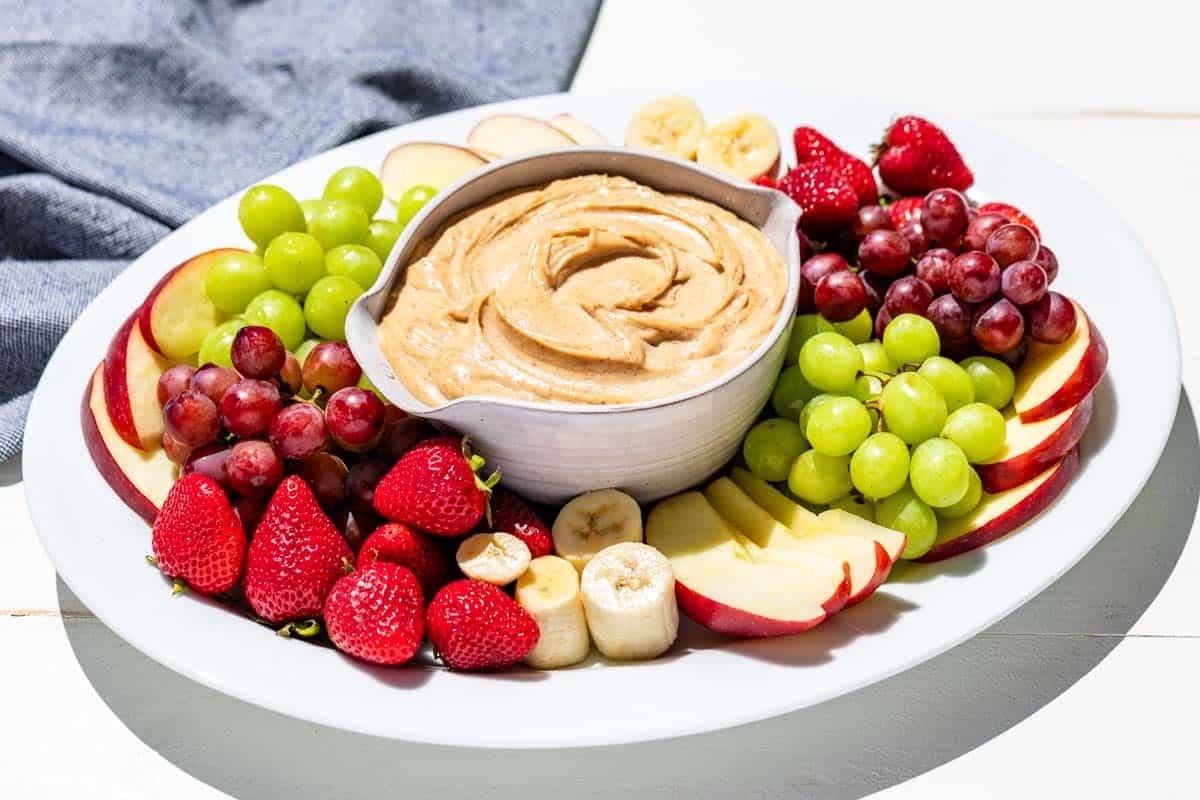 Finished peanut butter dip in a pottery bowl in the center of a large white platter surrounded by apple slices, grapes, strawberries, and banana slices.