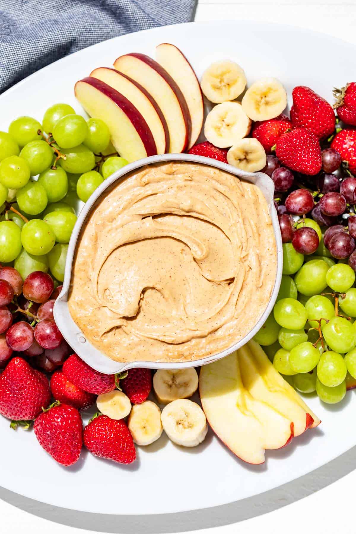 A large white platter with a bowl of peanut butter dip in the center surrounded by apple slices, banana slices, strawberries, and grapes.