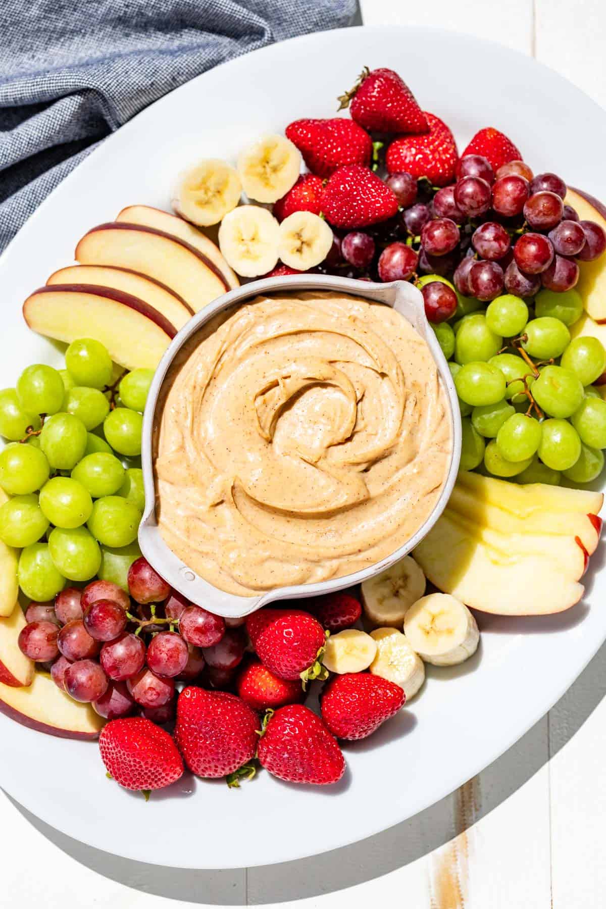 A large white platter with a bowl of peanut butter dip in the center surrounded by grapes, apples, banana slices, and strawberries.