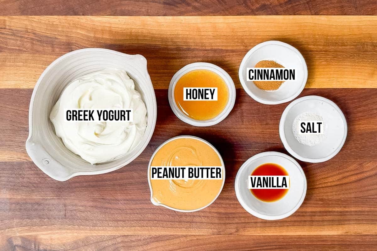 Bowls of Greek yogurt, peanut butter, honey, vanilla extract, cinnamon, and sea salt on a wood cutting board.