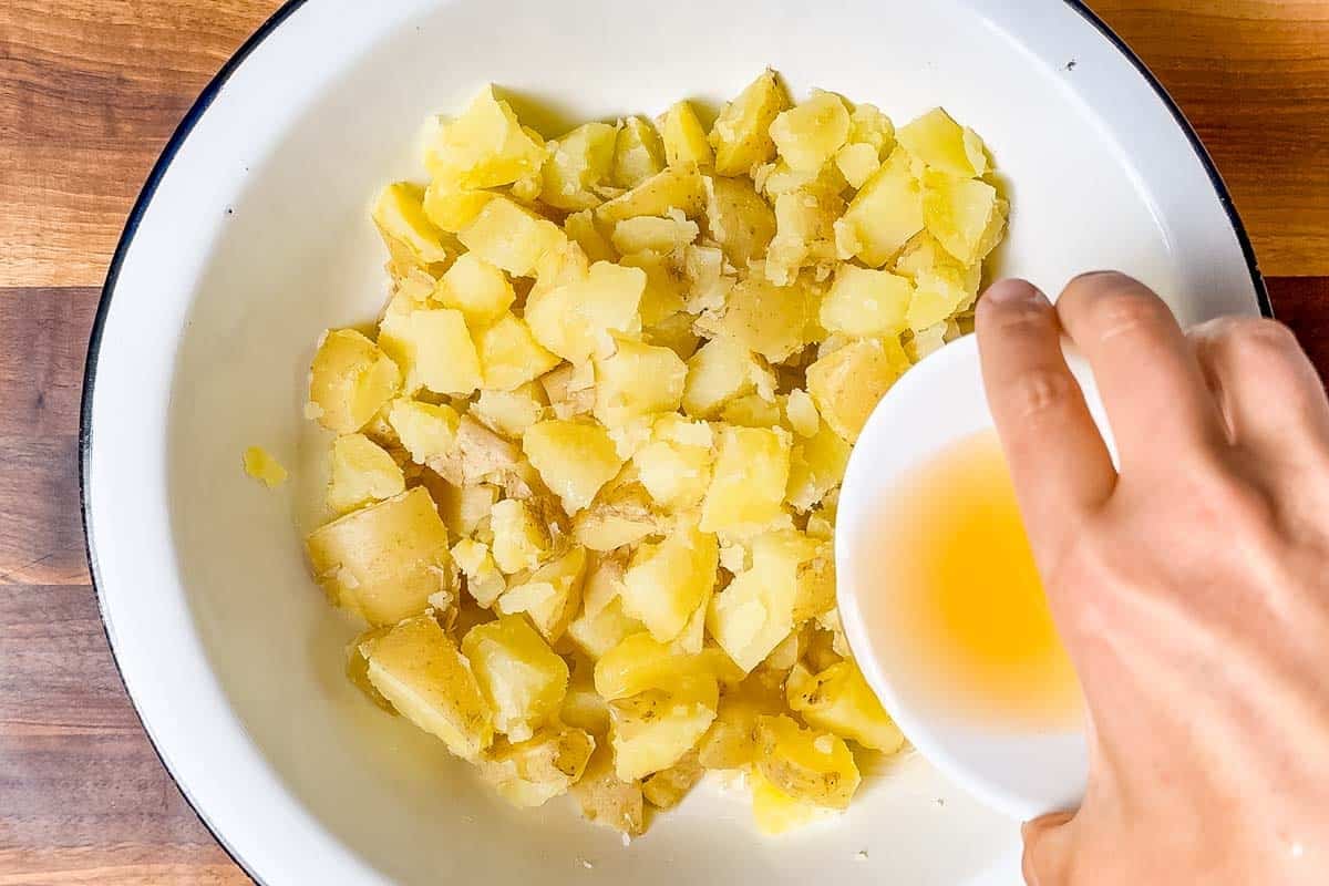 Pouring apple cider vinegar and pickle juice over the cut up potatoes in a large white bowl.