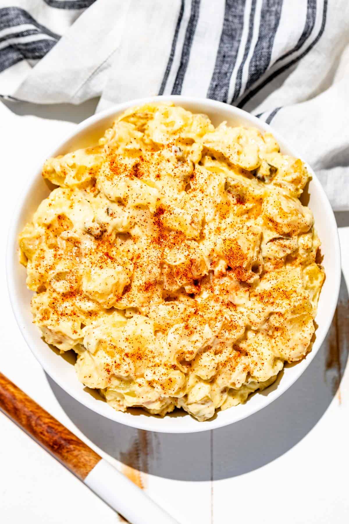 Potato salad topped with a sprinkle of paprika in a white bowl with a wood serving spoon on the side.