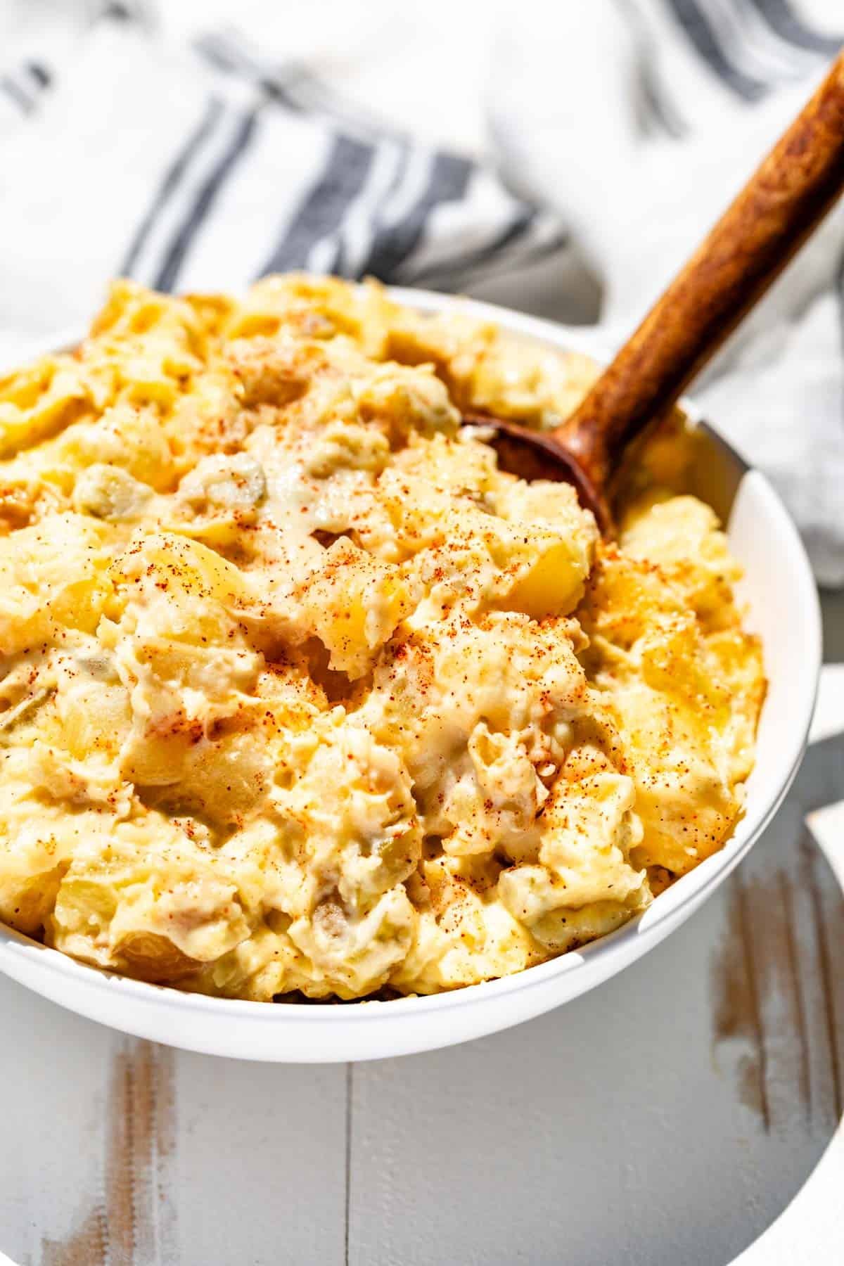 Potato salad in a white bowl with a wood serving spoon scooping some up.