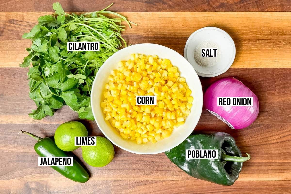 Ingredients for Roasted Chili Corn Salsa on a wood cutting board: corn, cilantro, jalapeno, limes, poblano, red onion. and sea salt.