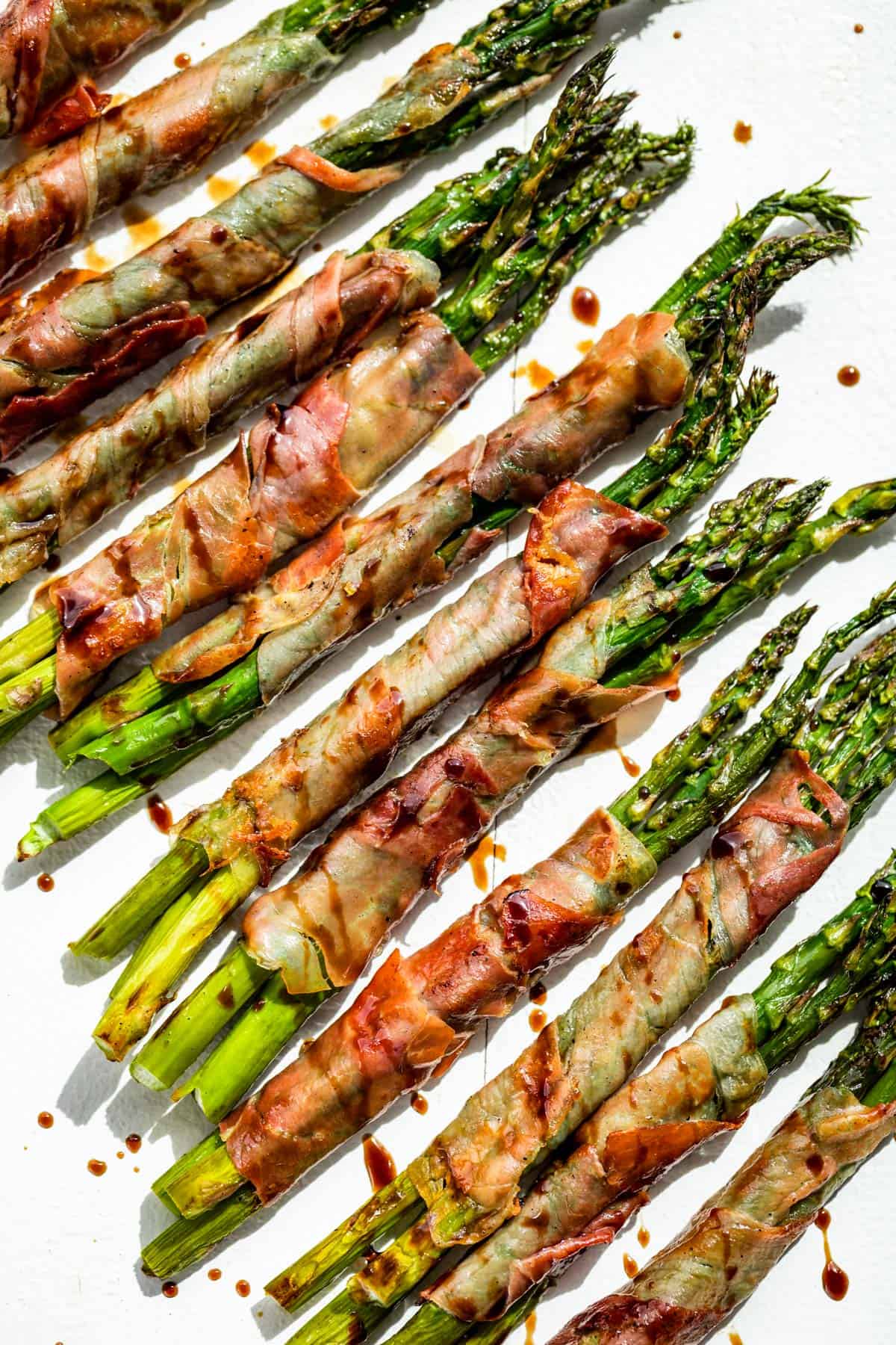 Prosciutto wrapped asparagus on a white board drizzled with balsamic vinegar.