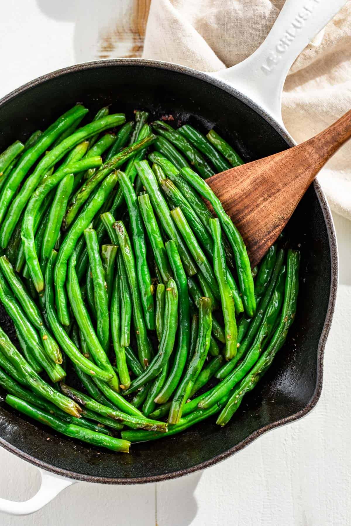 A large white cast iron skillet with green beans and a wood spatula.