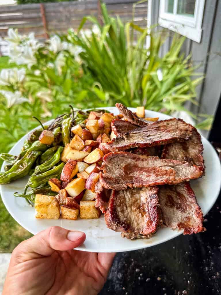 A white plate filled with shishito peppers, potatoes, and sliced tri tip with lilies and a green lawn in the background.