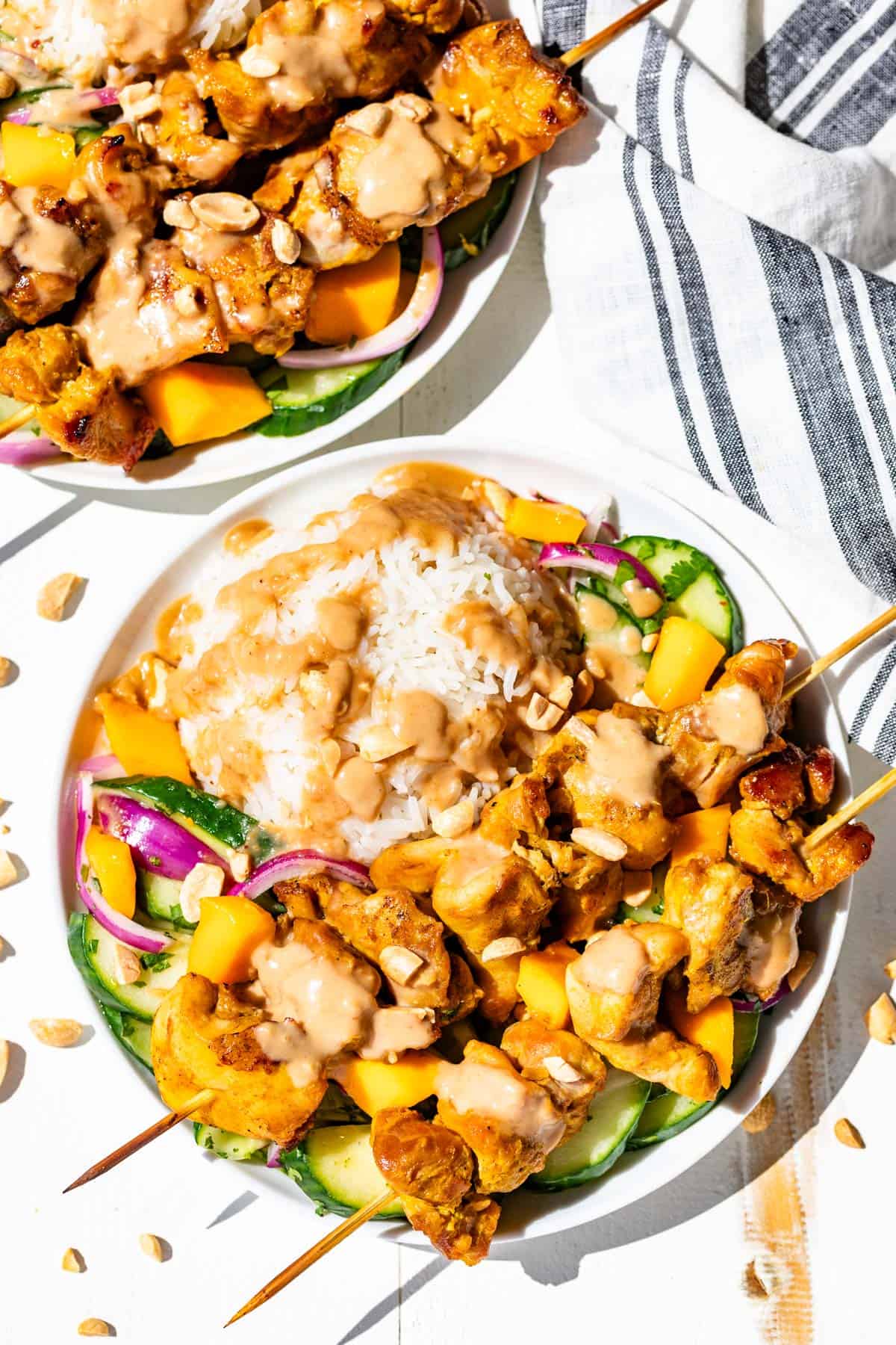 Two white plates with rice, Asian cucumber salad and turmeric chicken skewers drizzled with peanut sauce.