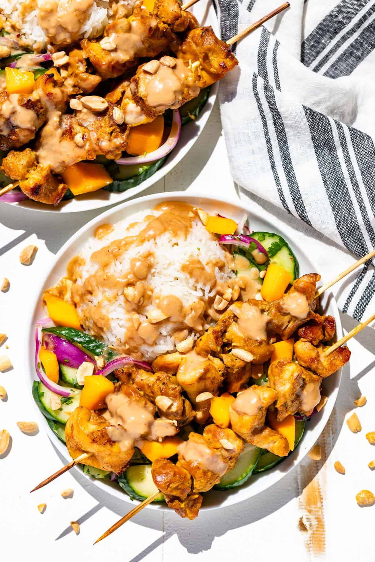 Two white plates with rice, Asian cucumber salad and turmeric chicken skewers drizzled with peanut sauce.