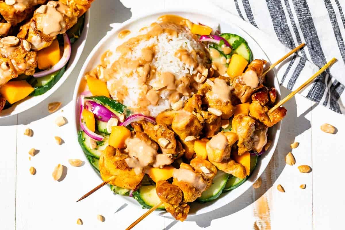 Finished turmeric chicken skewers sitting on top of Asian cucumber salad with rice on the side and drizzled with peanut sauce.