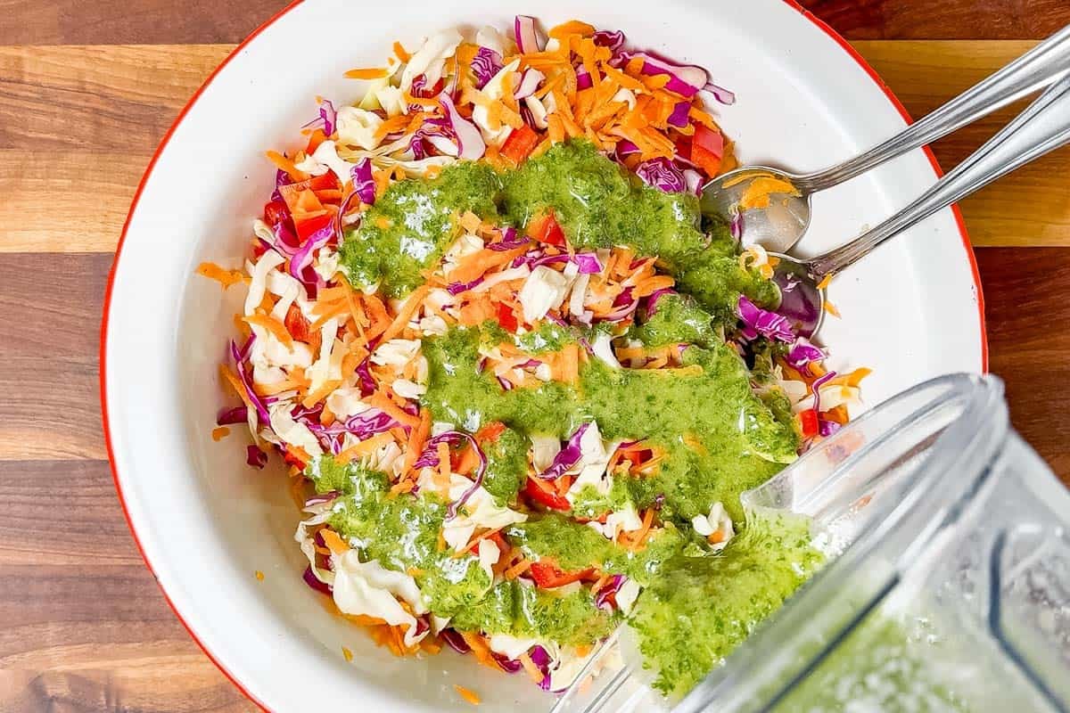 Pouring the cilantro lime dressing over the slaw mix in a large white bowl.