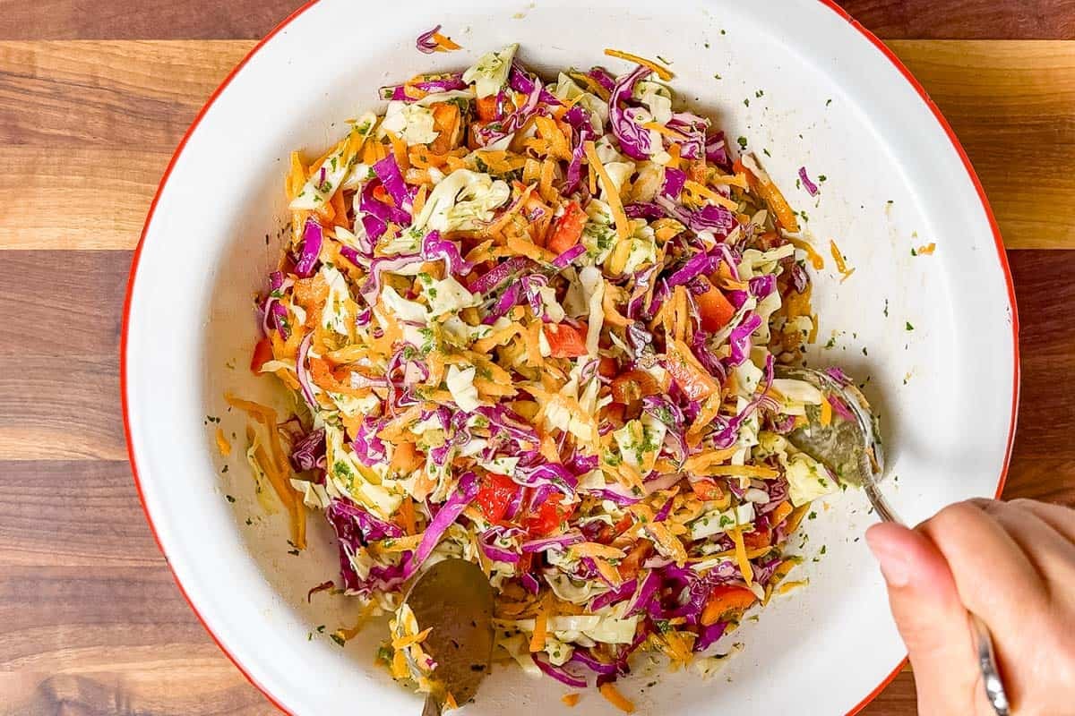 Tossing the slaw with the cilantro lime dressing with metal spoons in a large white bowl.