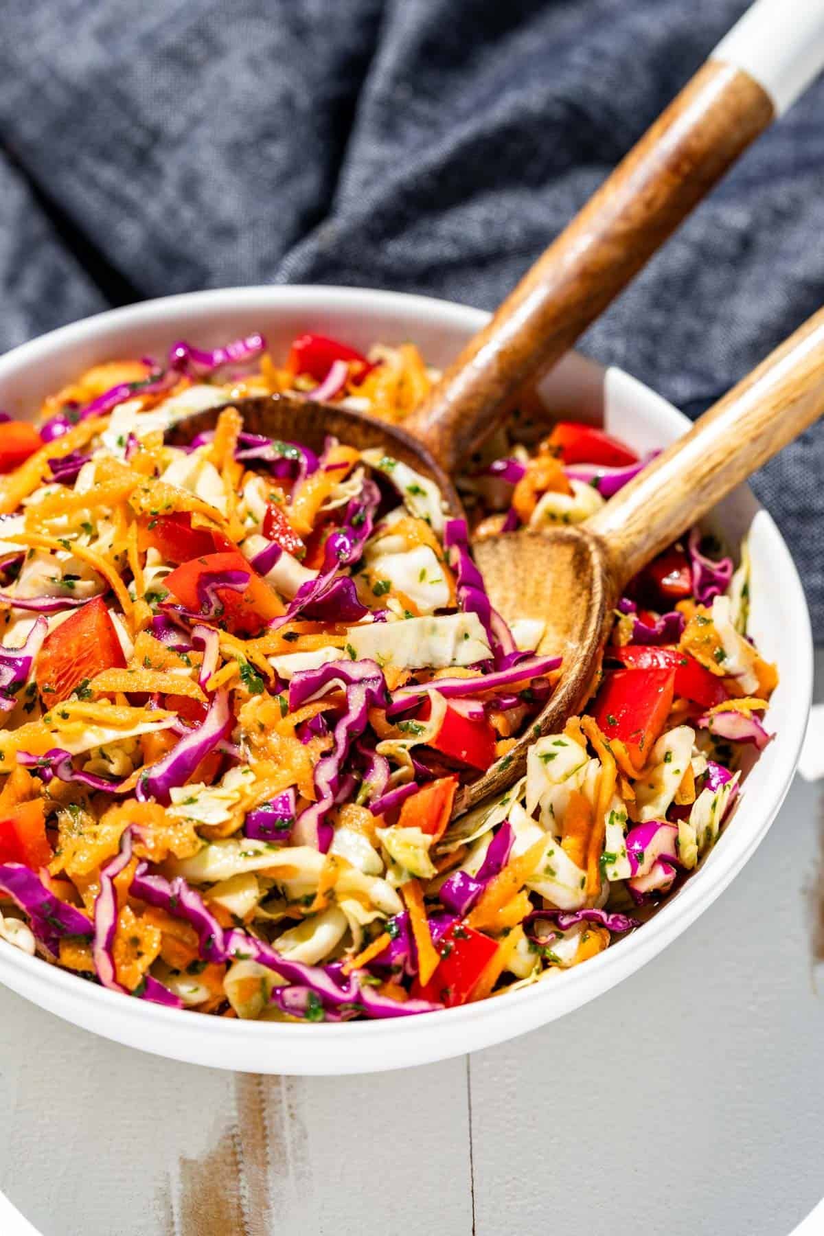 Cilantro lime slaw in a white bowl with wood serving spoons and a blue linen in the background.