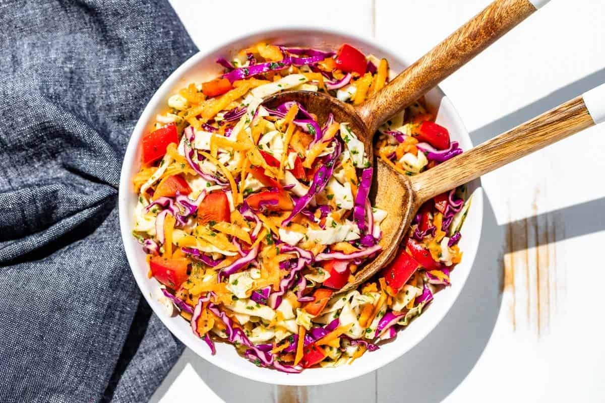 A large white serving bowl filled with Cilantro Lime Slaw and wooden serving spoons.
