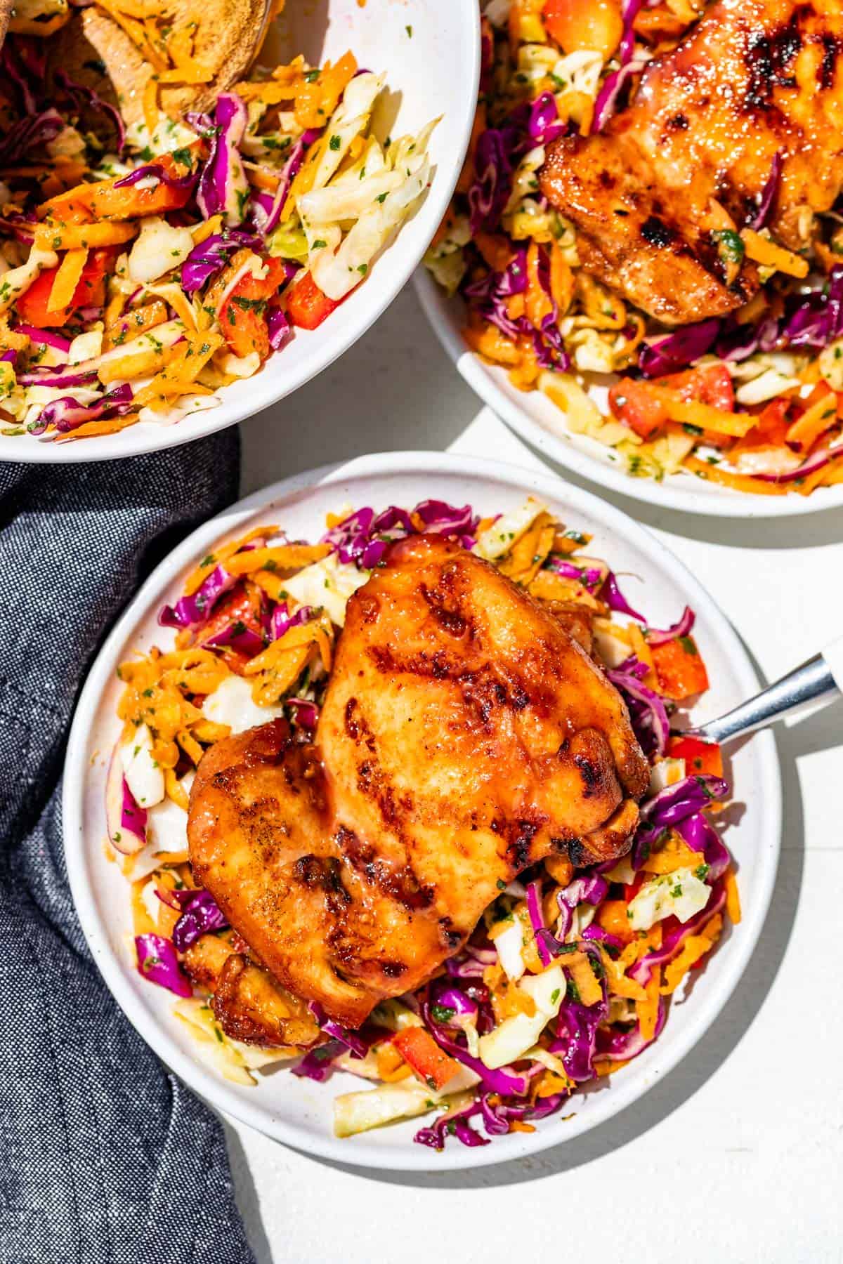 Two plates with cilantro lime slaw topped with honey sriracha chicken thighs with a bowl of slaw on the side.