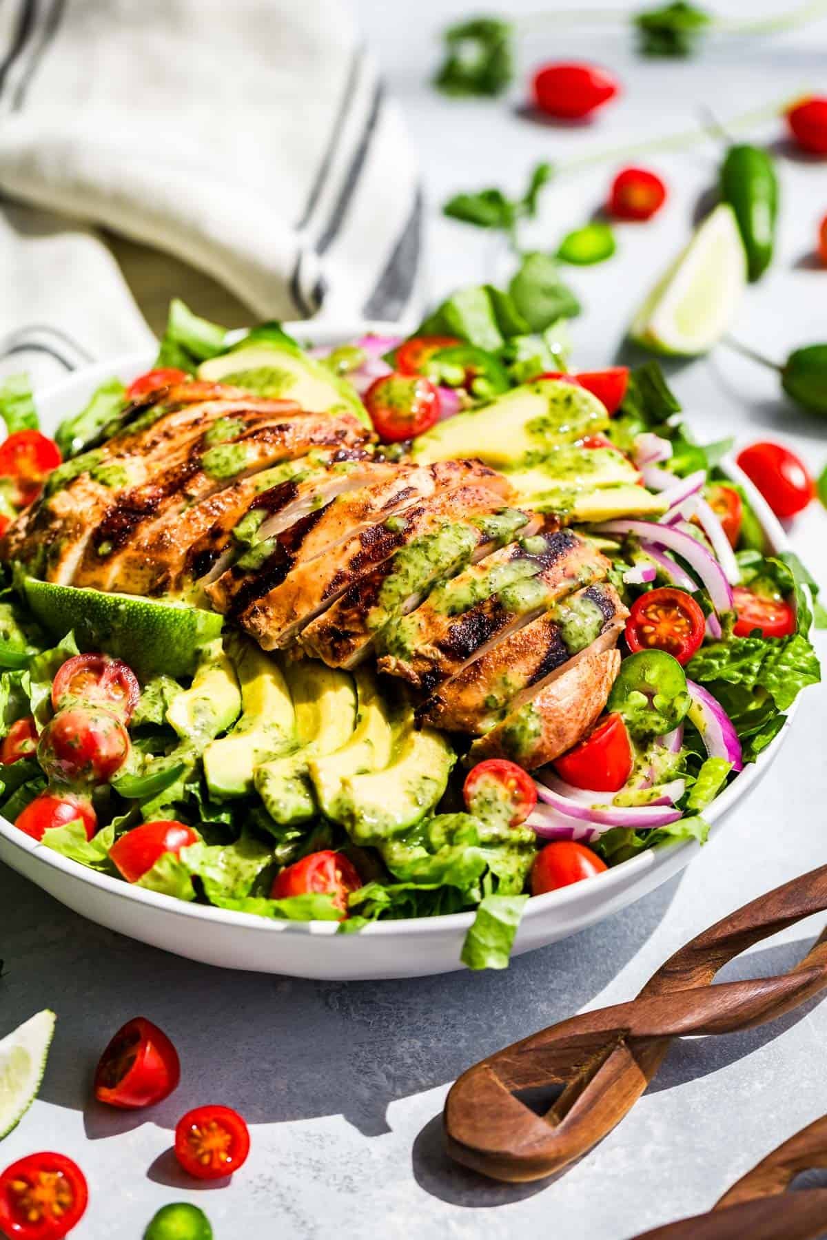 A large white bowl filled with guacamole salad topped with sliced avocado and sliced chipotle chicken with wood serving spoons on the side.