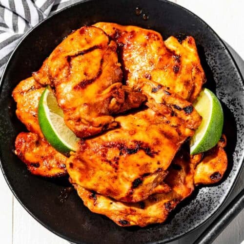 Honey Sriracha Chicken on a black plate with lime wedges with tongs and a striped linen on the side of the plates.