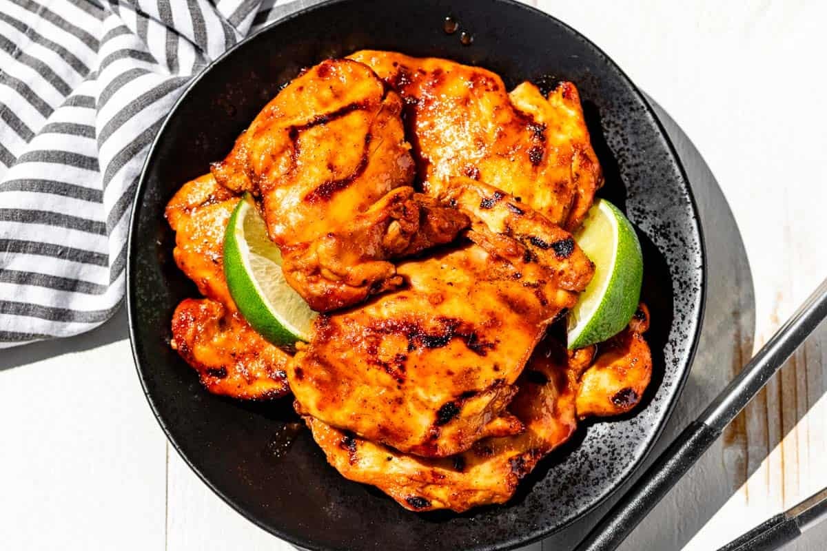 Honey Sriracha Chicken on a black plate with lime wedges with tongs and a striped linen on the side of the plates.