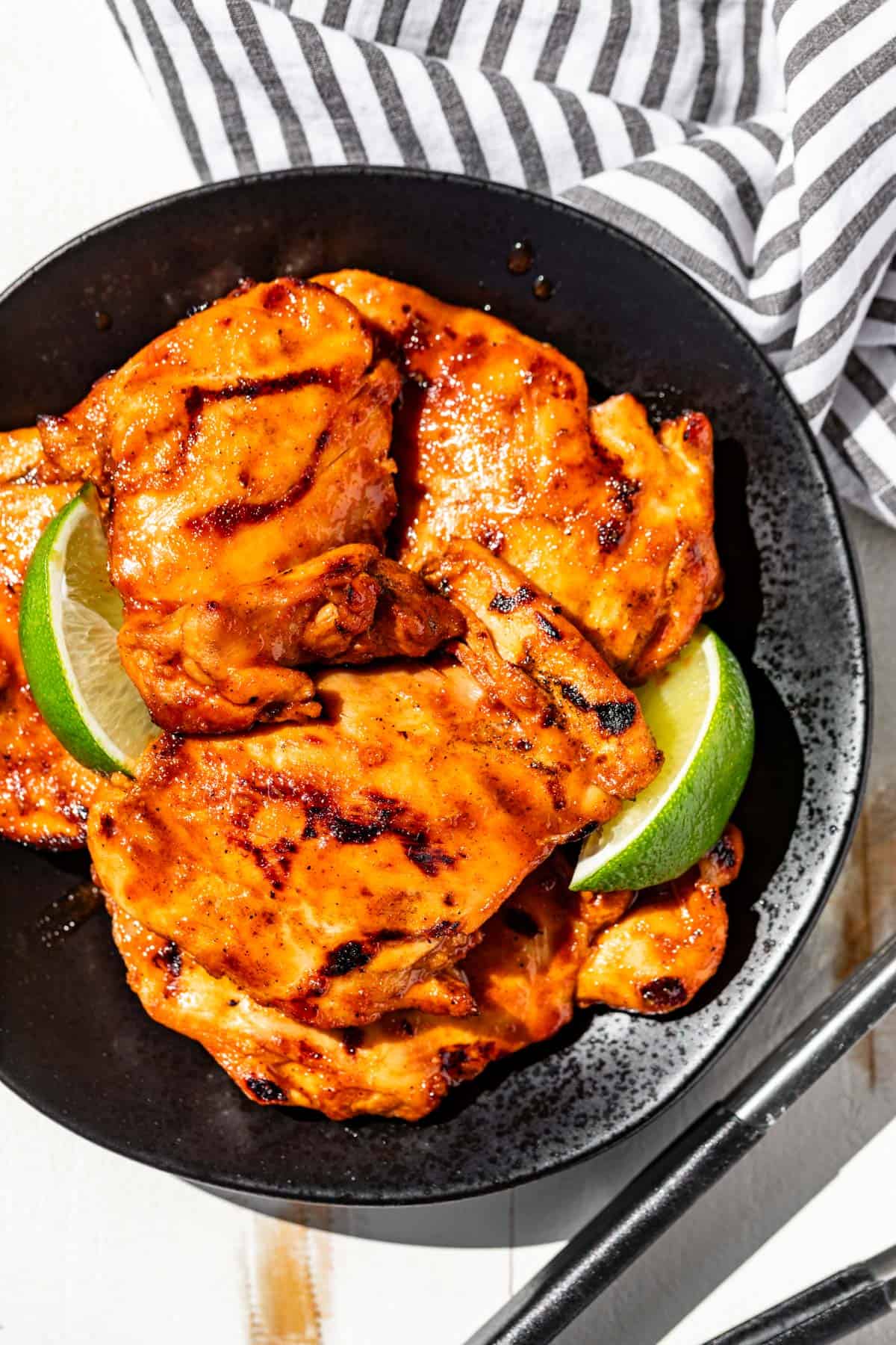 Honey Sriracha Chicken on a black plate topped with lime wedges and metal tongs on the side.