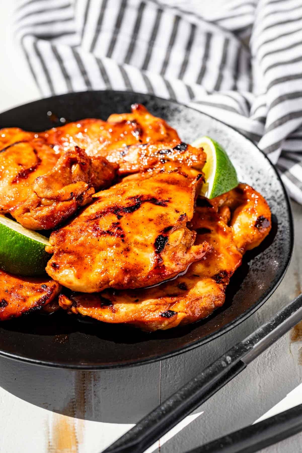 Honey Sriracha Chicken on a black plate with two lime wedges and tongs in front of the plate and a striped linen in the background.