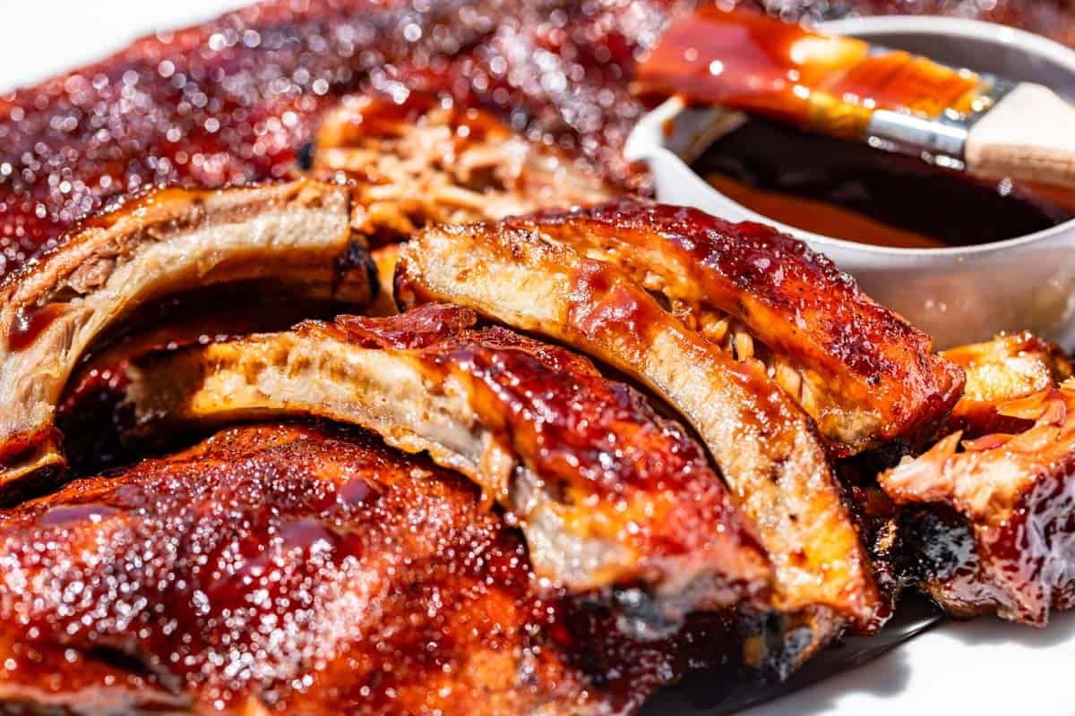 Sliced ribs with bbq sauce dripping over the edges and a bowl and brush with bbq sauce next to them.