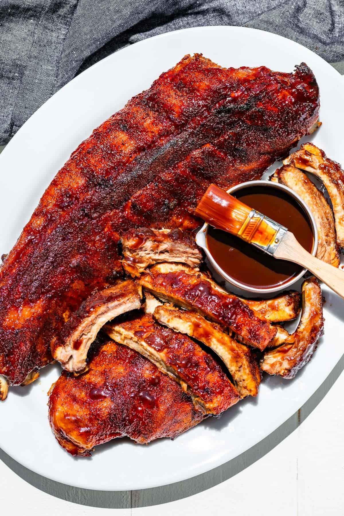 A large white platter with a full rack of baked bbq ribs and another rack of sliced bbq ribs next to it.
