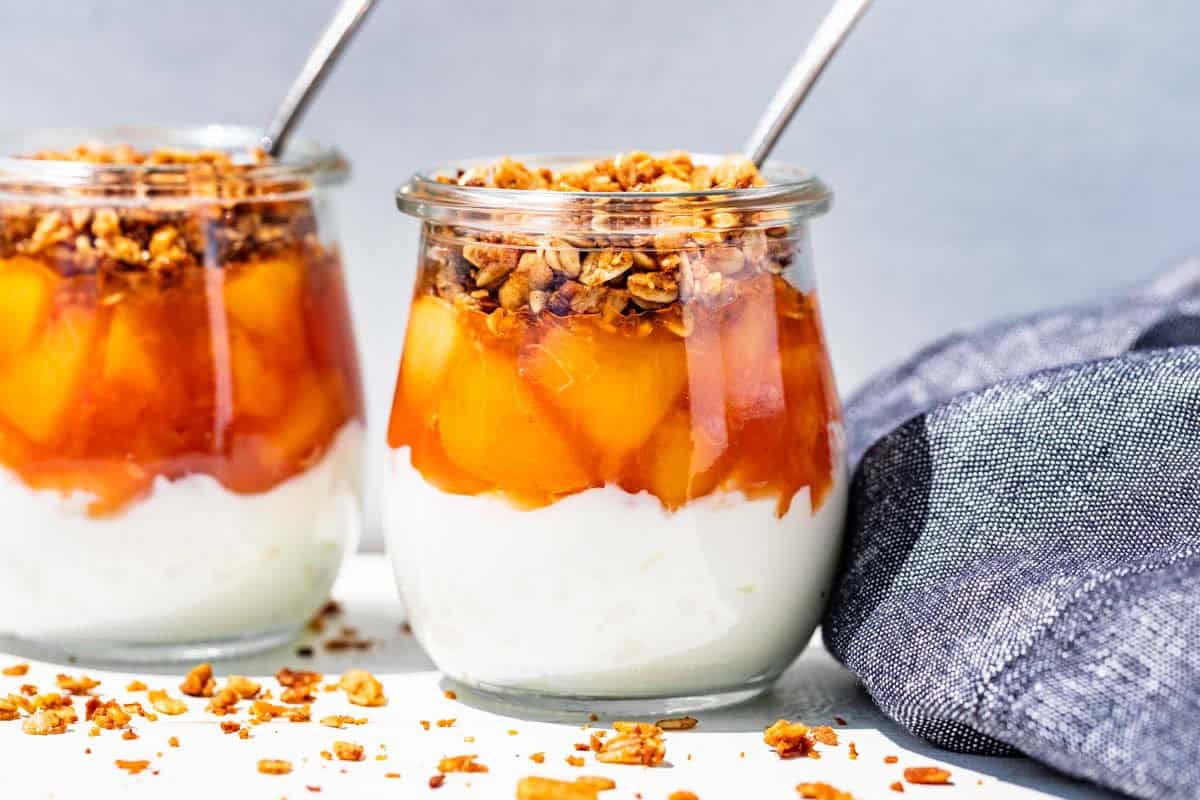 Peach sauce, yogurt, and granola layered into two glass jars with granola sprinkled around them.