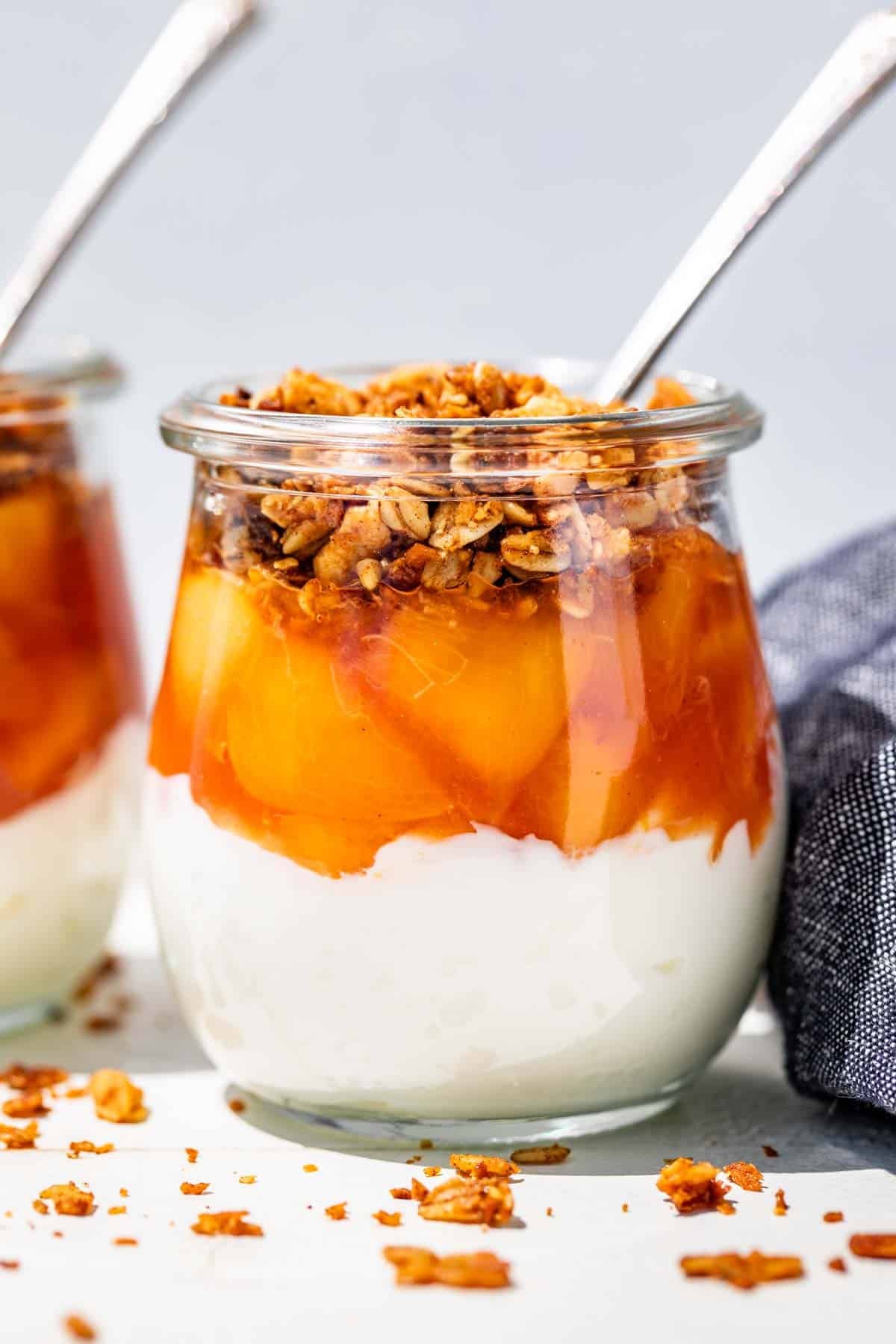 Two glass jars layered with yogurt, peach sauce, and granola with spoons in the jars.