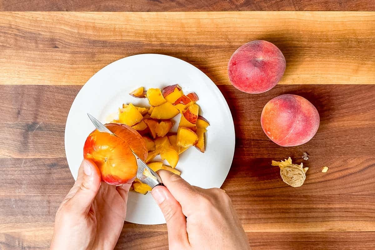 Cutting up a peach over a white plate on a wood cutting board with a paring knife.