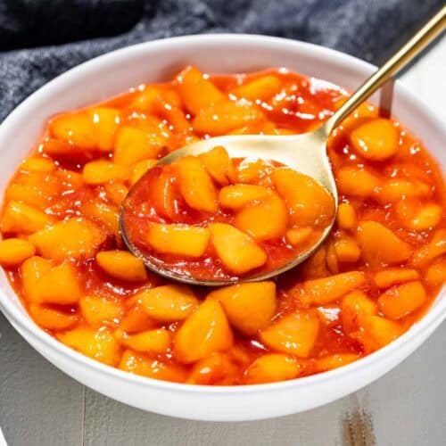 A white serving bowl filled with peach sauce with a gold spoon in it.