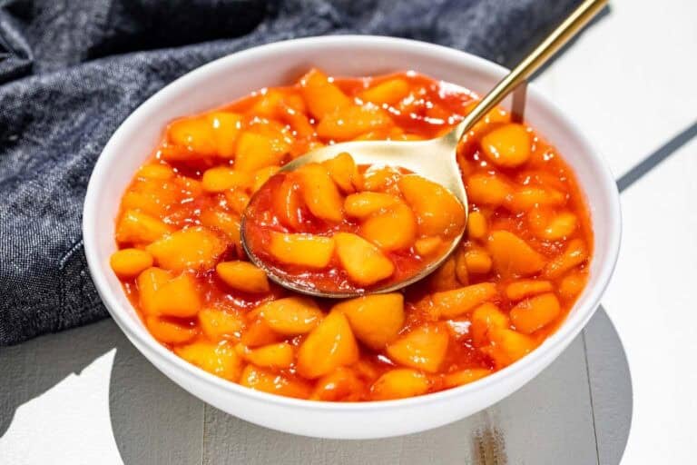A white serving bowl filled with peach sauce with a gold spoon in it.