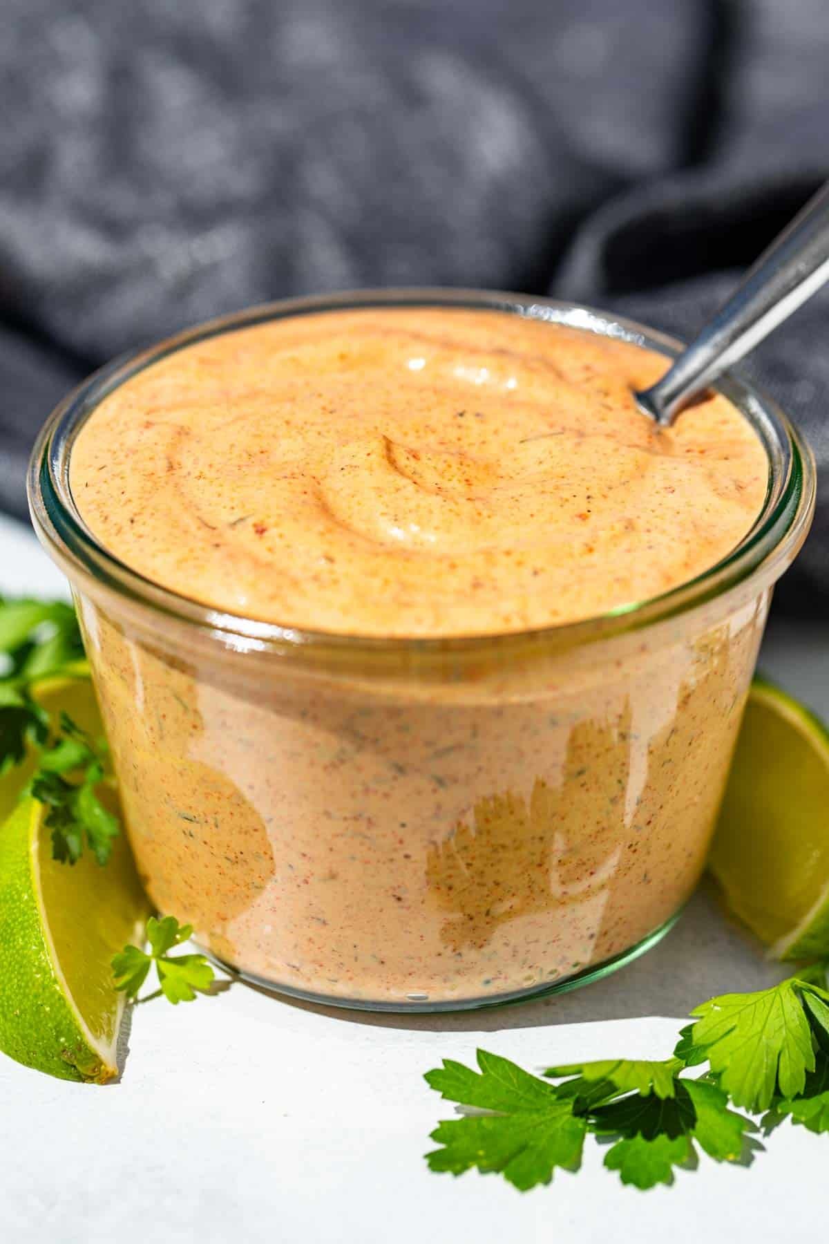 Chipotle ranch in a glass jar with a spoon in it with a blue linen in the background and lime wedges and cilantro around it.