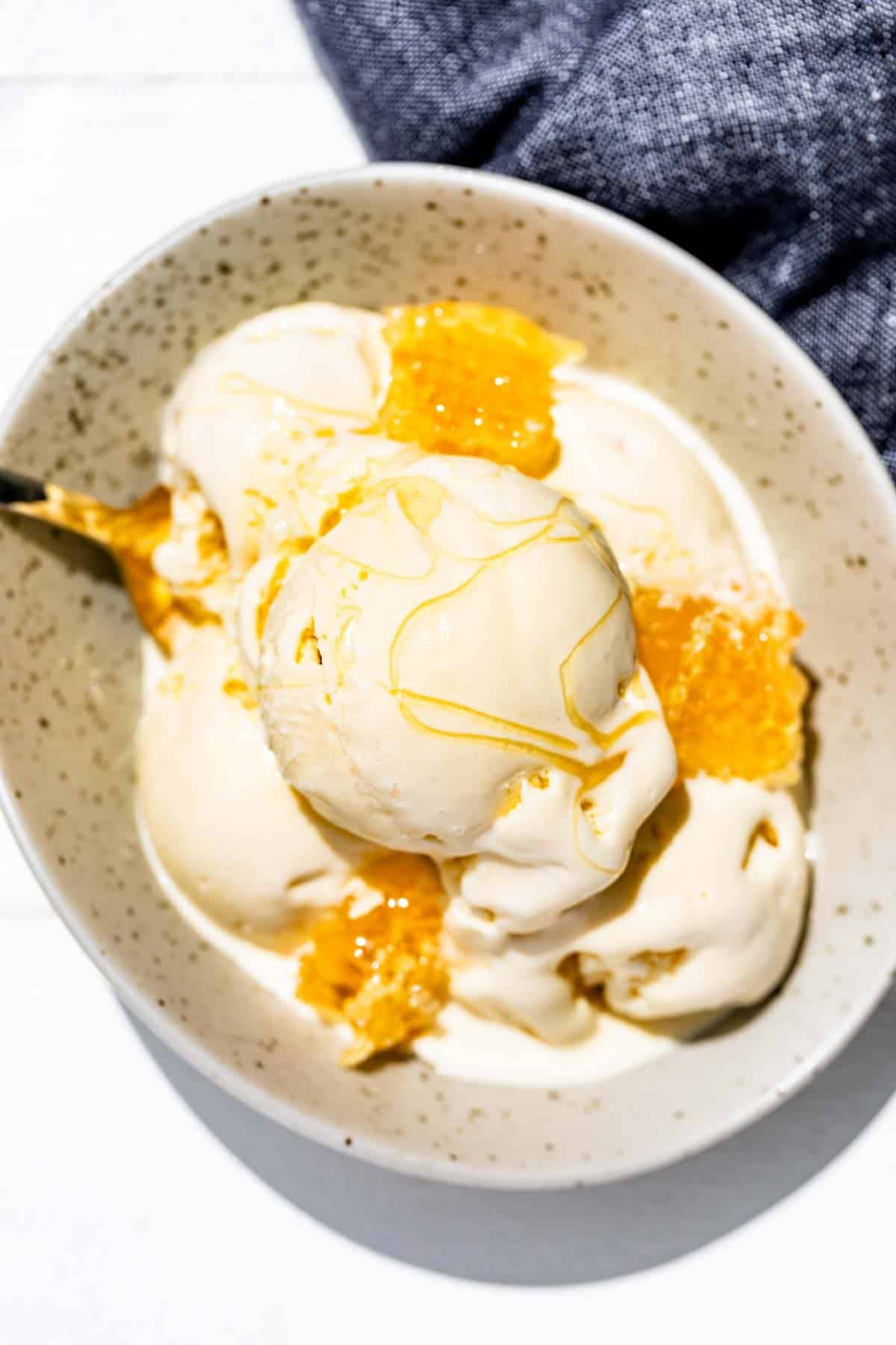 Five scoops of honey ice cream in a speckled pottery bowl with honeycomb and drizzled with honey.