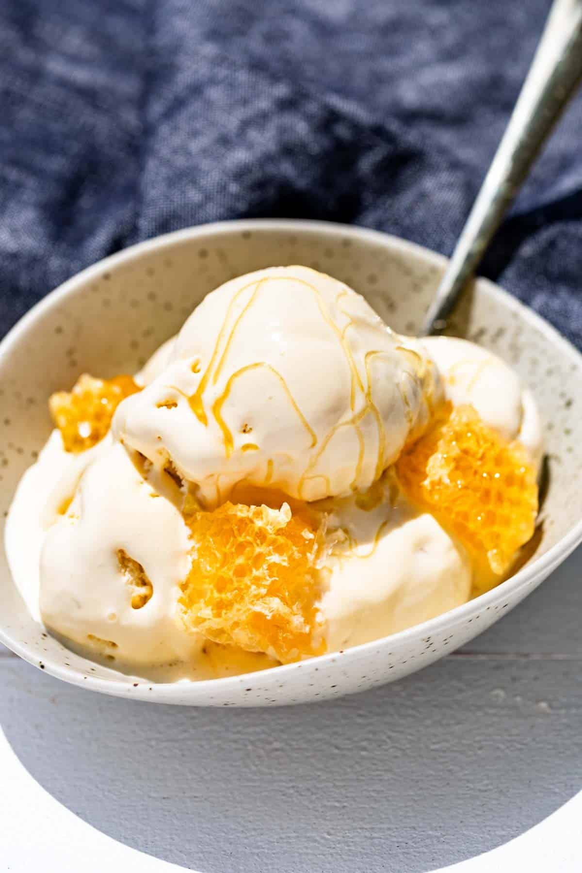 A pottery bowl filled with scoops of honey ice cream, honeycomb and drizzled with honey.