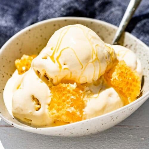 Four scoops of honey ice cream in a pottery bowl with a gold spoon, honeycomb and drizzled with honey.