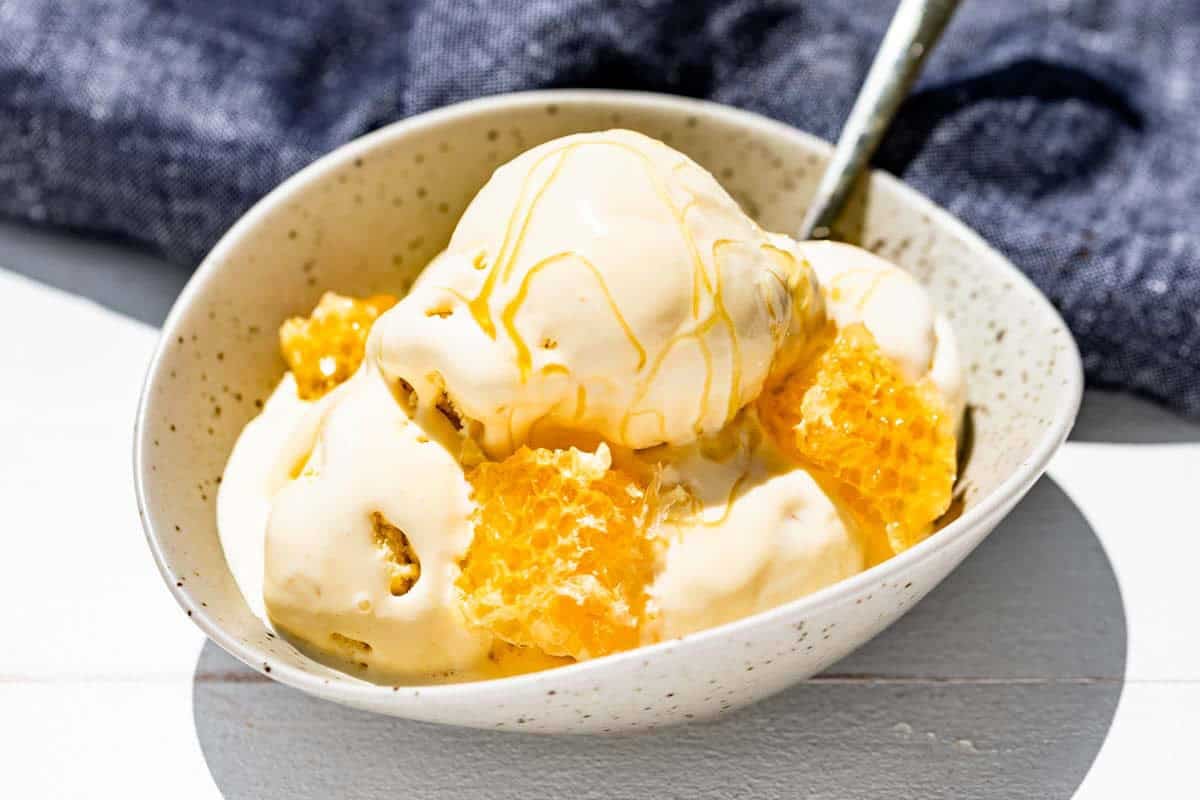 Four scoops of honey ice cream in a pottery bowl with a gold spoon, honeycomb and drizzled with honey.