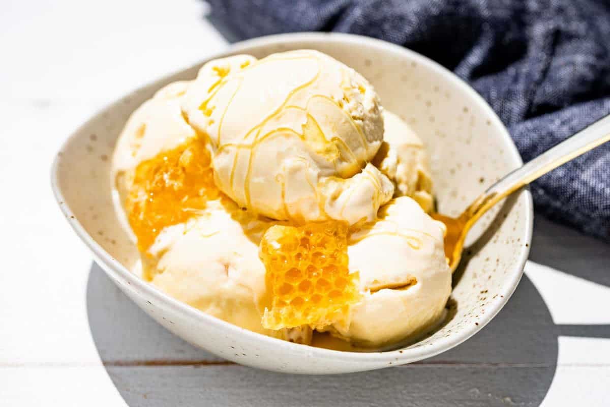 Scoops of honey ice cream in a pottery bowl with a gold spoon in it, chunks of honeycomb and drizzled with honey.