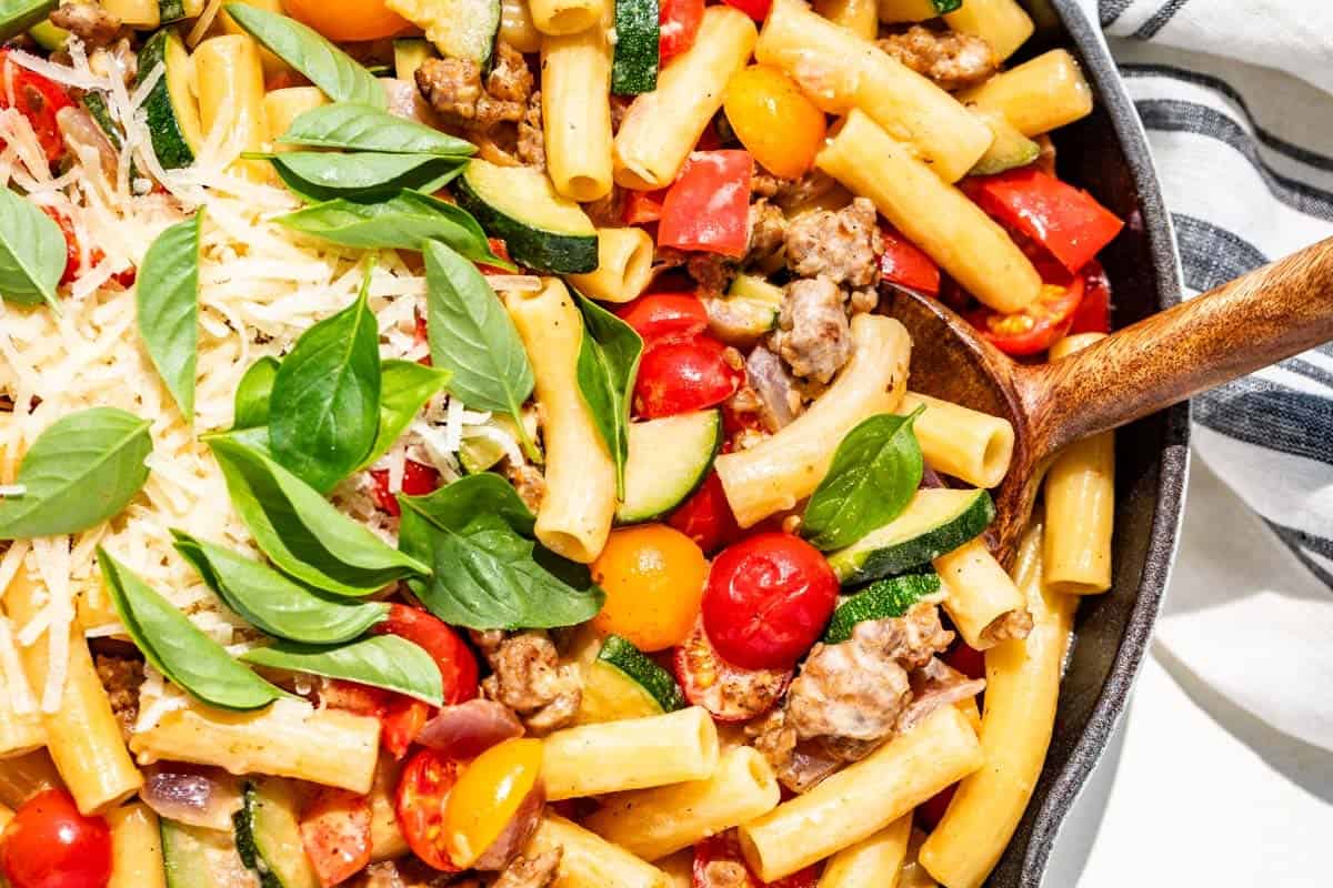 Sausage pepper pasta with zucchini, tomatoes, basil, and grated parmesan with a wood spoon.