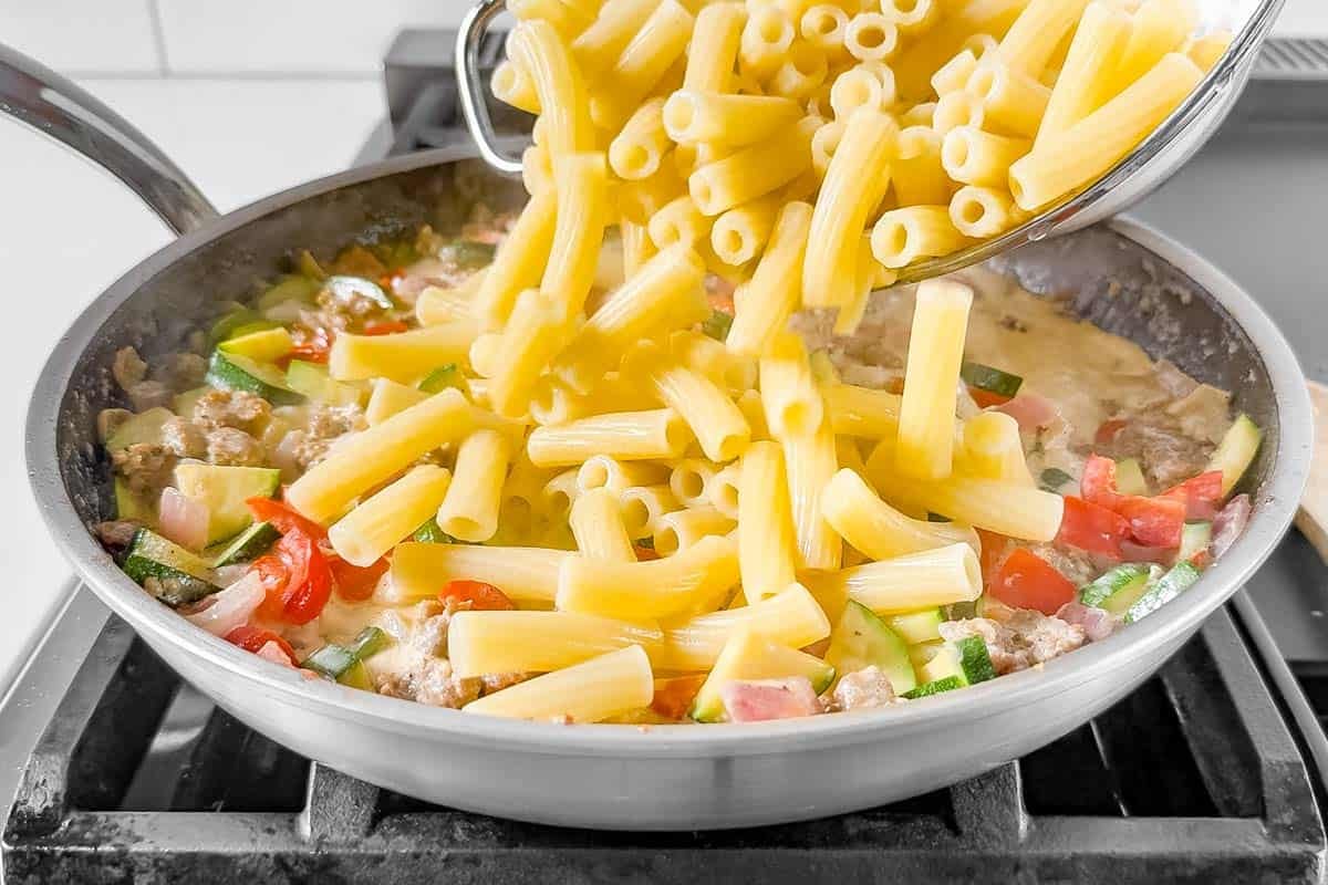 Adding the pasta to the sausage and peppers mixture in a large skillet on the stovetop.