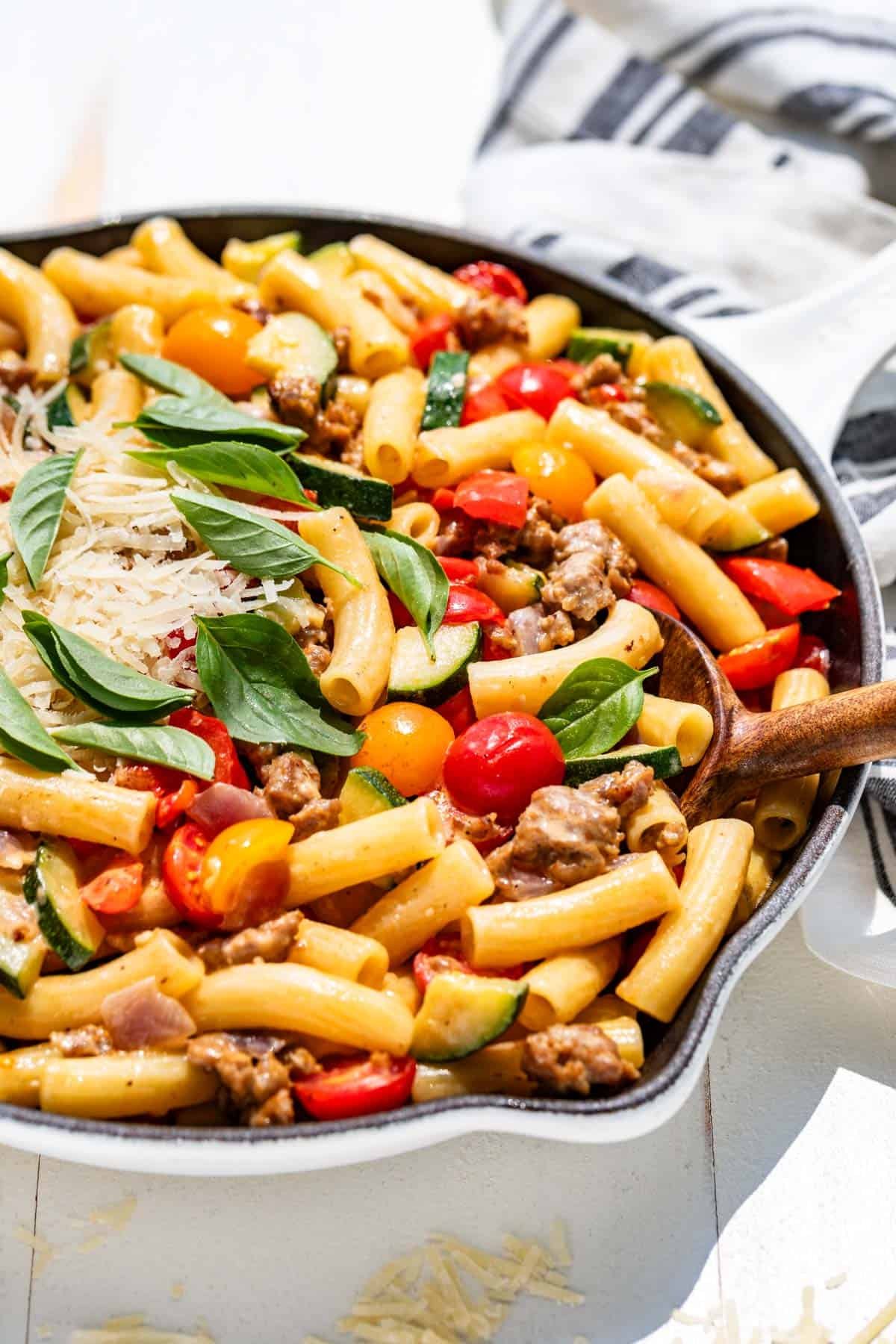 A white skillet filled with sausage and peppers pasta topped with grated parmesan and basil leaves.