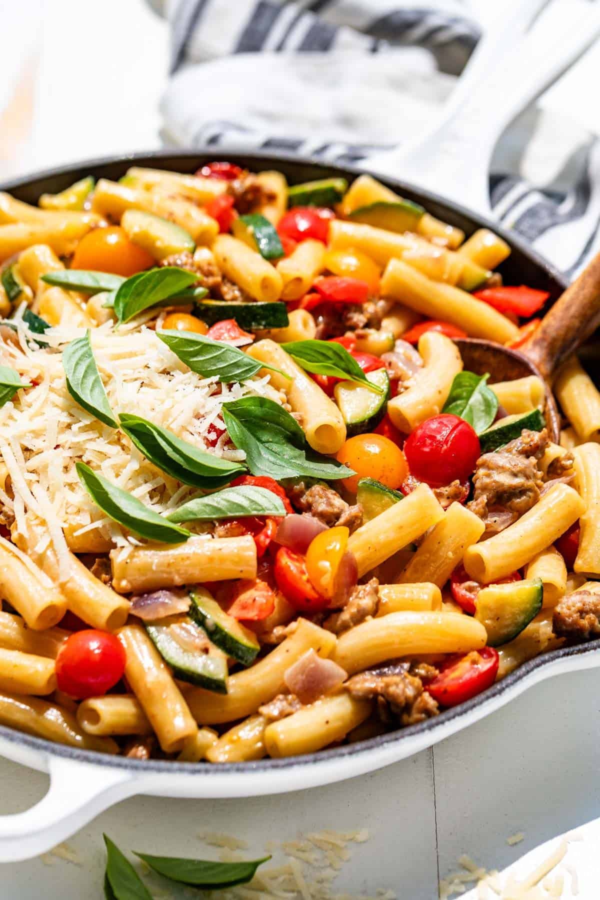 Sausage pepper pasta in a white skillet topped with grated parmesan and basil leaves with a wood spoon.
