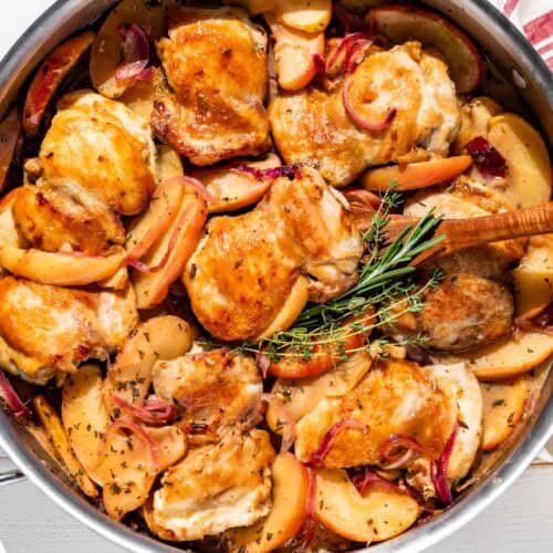 A large skillet with 8 chicken thighs surrounded by apple and onion slices, fresh rosemary and a wood spatula scooping one out.