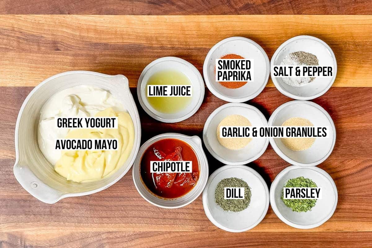 Bowls of mayo, Greek yogurt, chipotle in adobo, lime juice, dill, smoked paprika, dried parsley, garlic granules, and onion granules on a wood cutting board.