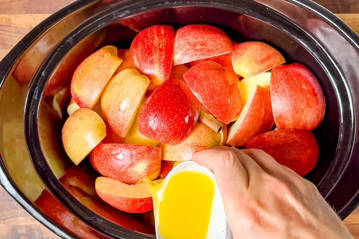 Adding the cinnamon stick and juice to the slow cooker bowl over the quartered apples.