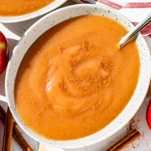 A pottery bowl filled with applesauce with a gold spoon in it with red apples and cinnamon sticks around the bowl.