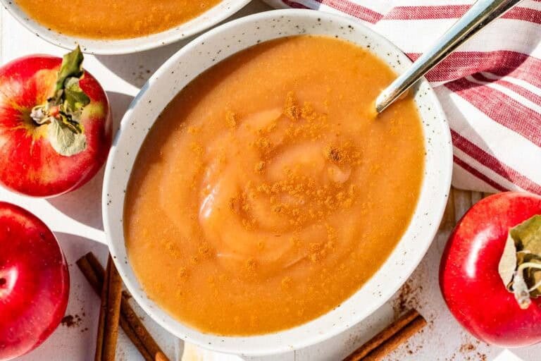 A pottery bowl filled with applesauce with a gold spoon in it with red apples and cinnamon sticks around the bowl.