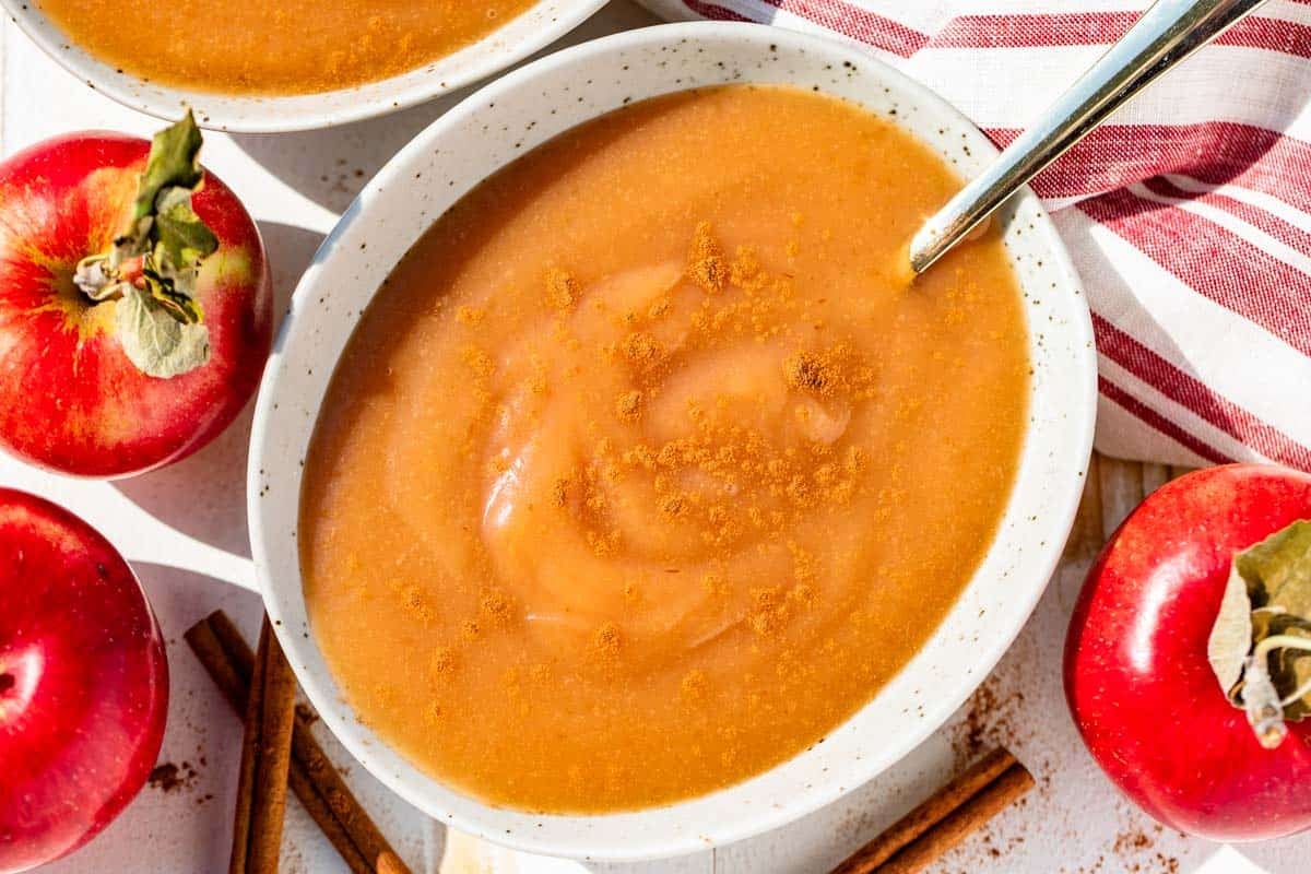 A pottery bowl filled with applesauce with a gold spoon in it with red apples and cinnamon sticks around the bowl.