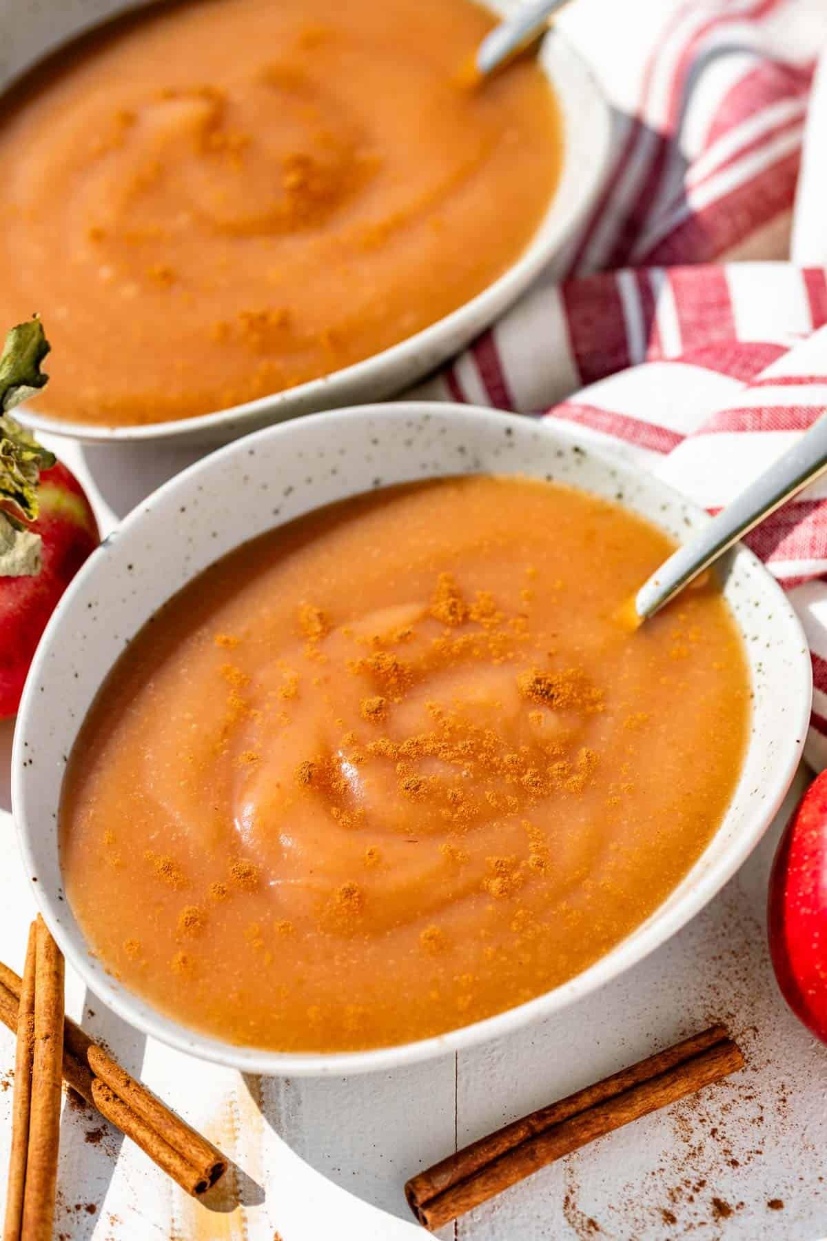 Two bowls of applesauce in pottery bowls sprinkled with cinnamon with gold spoons in the bowl and a red and white linen in the background.