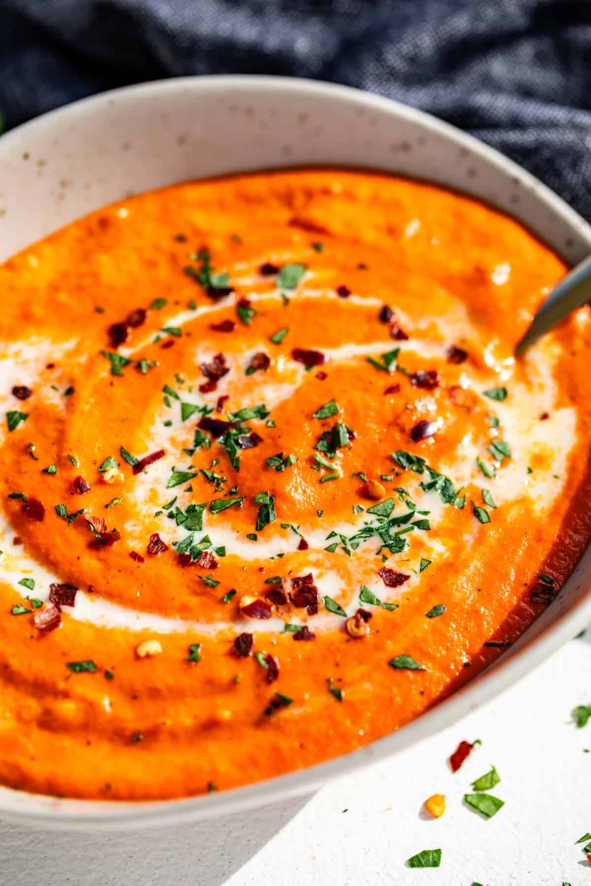 Roasted red pepper sauce in a pottery bowl topped with a drizzle of cream, topped pasley, and red pepper flakes with a gold spoon in it.